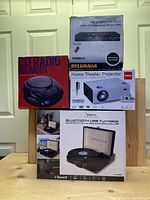 Overview of all four boxed items including CD Radio Boombox, HDMI Upconvert DVD Player, Sylvania Home Theater Projector, and Proscan Bluetooth USB Turntable.
