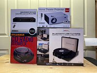 Overview of the four boxed electronic items: RCA Home Theater Projector, Proscan Bluetooth USB Turntable, Sylvania CD Radio Boombox, HDMI Upconvert DVD Player.