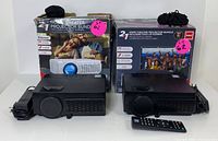 Two RCA home theater projectors, each with power adapter and remote control, alongside their respective boxed fold-up screens.