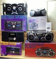 Photo showing Proscan Bluetooth CD Micro System, Proscan Bluetooth CD Boombox, Proscan Radio CD/Cassette Boombox, and Bluetooth speaker system with packaging and remote included.