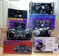 Photo showing entire bundle with multiple boxed and unboxed audio devices including Proscan CD Micro System box, Bluetooth speakers, CD Bluetooth Boombox, and Crosley Radio CD/Cassette Boombox with headphones.