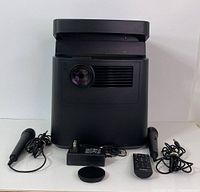Front view of the RCA 2-in-1 portable projector with two wired microphones, power adapter, remote control, and lens cap laid out.