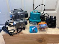 Photo showing the two silver grey Vivosun air pumps, the turquoise Vivosun submersible pump with handle, the black Aquaccom submersible pump, and two Robo Fish toys in plastic cases (blue and orange clownfish).