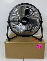 Front view of the Deluxe high-velocity floor fan showing metal blades and black metal cage, sitting on a cardboard box.
