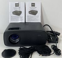 Front view of RCA RPJ714 black multimedia projector with lens cap removed, power cable, user manuals placed behind the projector.