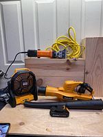 Cordless leaf blower, portable fan, battery charger with two batteries, electric sheep shears with yellow cord on wooden surface.