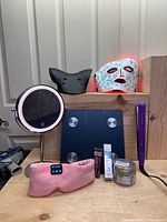 Photo showing LED vanity mirror, purple hair straightener, pink neck/shoulder relaxer, body scale, cosmetic items including tattoo brow tint, dark spot removal cream, aloe vera gel, and Bluetooth face mask on shelf
