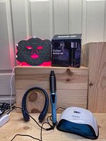 Five beauty and wellness devices arranged on a wooden surface: a black LED light therapy mask lit pink, Gvber massage gun box, curling iron brush, neck fan, UV nail light with display.