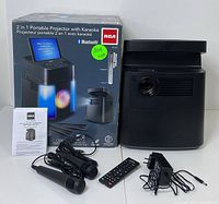 Full view of RCA 2-in-1 portable projector with karaoke system including original box, two wired microphones, power adapter, remote control, and instruction manual.