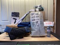 Full lot view including couch cover in blue fabric, Shiatsu massager, electric scrub brush attachments in box, handheld steamer, cordless vacuum, and soft pillow covers in plastic bag.