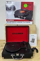 Sylvania Bluetooth USB/SD turntable in black suitcase with red interior. Includes power cord and manual. Box shown in background.