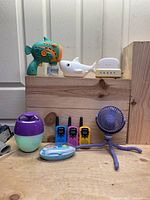 Items displayed on wooden surface including waterproof floating speaker, electric nail grinder, walkie talkies, night lights, portable fan, and fish bubble machine