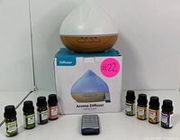 Photo showing the aroma diffuser placed on top of its original box with seven essential oil bottles arranged on both sides, and a black remote control in front.