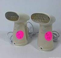 Front view of two white handheld garment steamers with multi-hole beige steam plates and power cords coiled beside them, each marked with pink stickers.