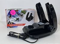 Electric black plastic footwear dryer with foldable arms and attached power cord, displayed with original packaging box.