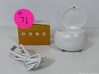 Photo showing white oval-shaped hearing aid dry box with lid open, power button on front, and included USB charging cable.