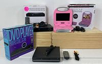 Overview of electronics package showing pink portable DVD player in open position with box, black standard DVD player, remote controls, power and AV cables, and digital converter box.