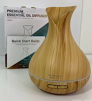 Photo shows the diffuser placed in front of its original box and quick start guide. The diffuser has a natural wood grain pattern with control buttons on the front.