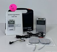 TENS 7000 device with instruction manual, electrode pads, cables, and black hard case arranged on white background.