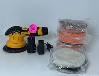 Yellow and black cordless car polisher with charger and stack of buffing pads wrapped in plastic on white background