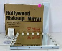 Hollywood makeup mirror flat on floor showing 15 round LED bulbs around perimeter and white side panel. Original cardboard box and accessories visible behind.