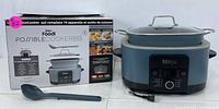 Ninja Foodi Possible Cooker Pro multi-cooker with box, cooking pot with glass lid on top, and blue-gray plastic serving spoon in front.