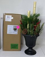 Photo showing the black urn filled with greenery, red berries, pinecones, and two gold-tone candles, with original packaging box beside it.