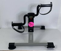 Full view of the compact pedal exerciser showing gray metal base and black pedal arms with foot straps, and central LCD display unit.