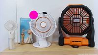 Set of three fans on wooden surface, industrial fan on right, desktop fan center, handheld spray fan left with packaging visible behind