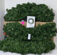 Photo showing three lush green pre-lit Christmas garlands stacked together with a remote and a product manual booklet.