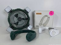 Dark green rechargeable fan with LED lights next to white portable neck fan and its box on a white background
