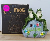 Front view of the solar-powered frog welcome statue showing two green frogs on a welcome stone with colorful flowers and original packaging box in background.