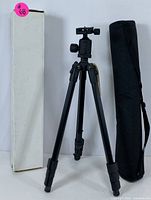 Black camera tripod fully extended with flip-lock legs, ball head mount visible. Positioned between a white box and black carrying case.