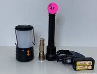 Outdoor lighting set featuring a black rechargeable LED camping lantern with frosted diffuser, a tall black flashlight, a compact metallic gold flashlight, an Everbeam headlamp, and rechargeable AA batteries with charger.