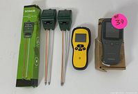 Four measuring devices on a white surface including two green soil meters with metal probes, a yellow digital stud finder, and a black IGEREES multifunction air detector on its box.