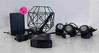 Photo showing entire lot items from a side angle: 3 LED solar spotlights with cables, decorative black metal cage light, small solar panel charger, outdoor power extension with 3 outlets and cord.
