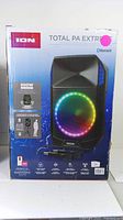 Front view of boxed ION Total PA Extreme speaker system showing branding, 600W output, Bluetooth logo, wireless mic image, and LED light ring on speaker.