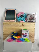 Display of all toys included: pink LCD writing tablet, blue and pink toy steering wheel, boxed puzzle game, flying spinner set including launcher gun and six colorful discs arranged on a white rear surface with wooden backdrop.