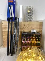 Shows the black adjustable curtain rods (3 pieces), star curtain lights with LED stars lit in warm yellow, a pack of button cell batteries, USB charger, and a wooden board background.