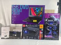 Wide photo showing all four main items with their packaging: RCA projector box, portable DVD player box, Bluetooth CD micro system box, and Sylvania Bluetooth speaker box.