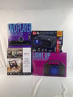 Photo of four boxed items in the lot: a DVD player with progressive scan, RCA HD home theatre projector with bonus screen, light up Bluetooth speaker, and 9" dual screen portable DVD player.