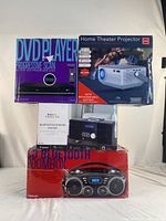 Photo showing boxed DVD player with progressive scan technology, RCA Home Theatre Projector box, Bluetooth CD micro system box, and CD Bluetooth boombox box stacked