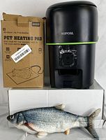Photo showing the covered automatic pet feeder with front camera, dog heating pad box, and a realistic fish toy.