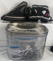 Pet water fountain in stainless steel with clear reservoir and filtration system, three black rabbit harnesses with padded mesh and reflective trim on white background.