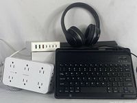 All items displayed together: wireless keyboard tablet case black, wireless headphones black, white power bar with six outlets, and black USB hub with multiple ports