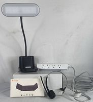 LED desk lamp with adjustable neck and storage base next to white power bar with multiple outlets and a coiled power cord. Wireless keyboard box and black wireless headset with a microphone in front of them.