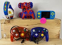 Photo showing two GameCube-style wired controllers, red and blue, RC stunt car in red and blue, handheld gaming console blue with yellow controls, and Super Mario-themed headphones