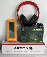 Photo of red and black over-ear headphones, orange box labeled 'ARGB FAN HUB' containing 9 port ARGB fan hub, 2.4G wireless gaming controller box, and ArgonX Helix small gaming mouse pad in white box.