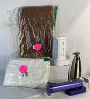 Five items displayed on white surface: brown bathroom mats in packaging, white pants in packaging, purple garment steamer with charging base, white tower power strip, stainless steel soap dispenser.