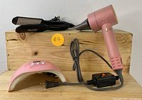 Photo showing all three items: pink hairdryer, black hair crimper, and pink LED nail lamp positioned on wooden surface.